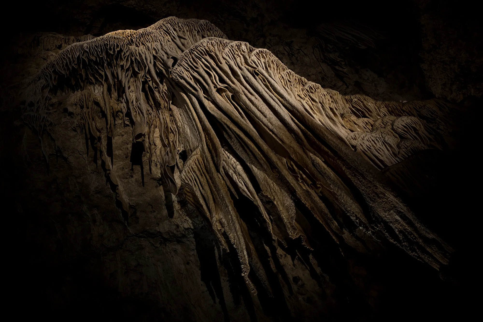 Rippled sheets of pale cave flowstone hanging in folds, lit from one side against deep darkness