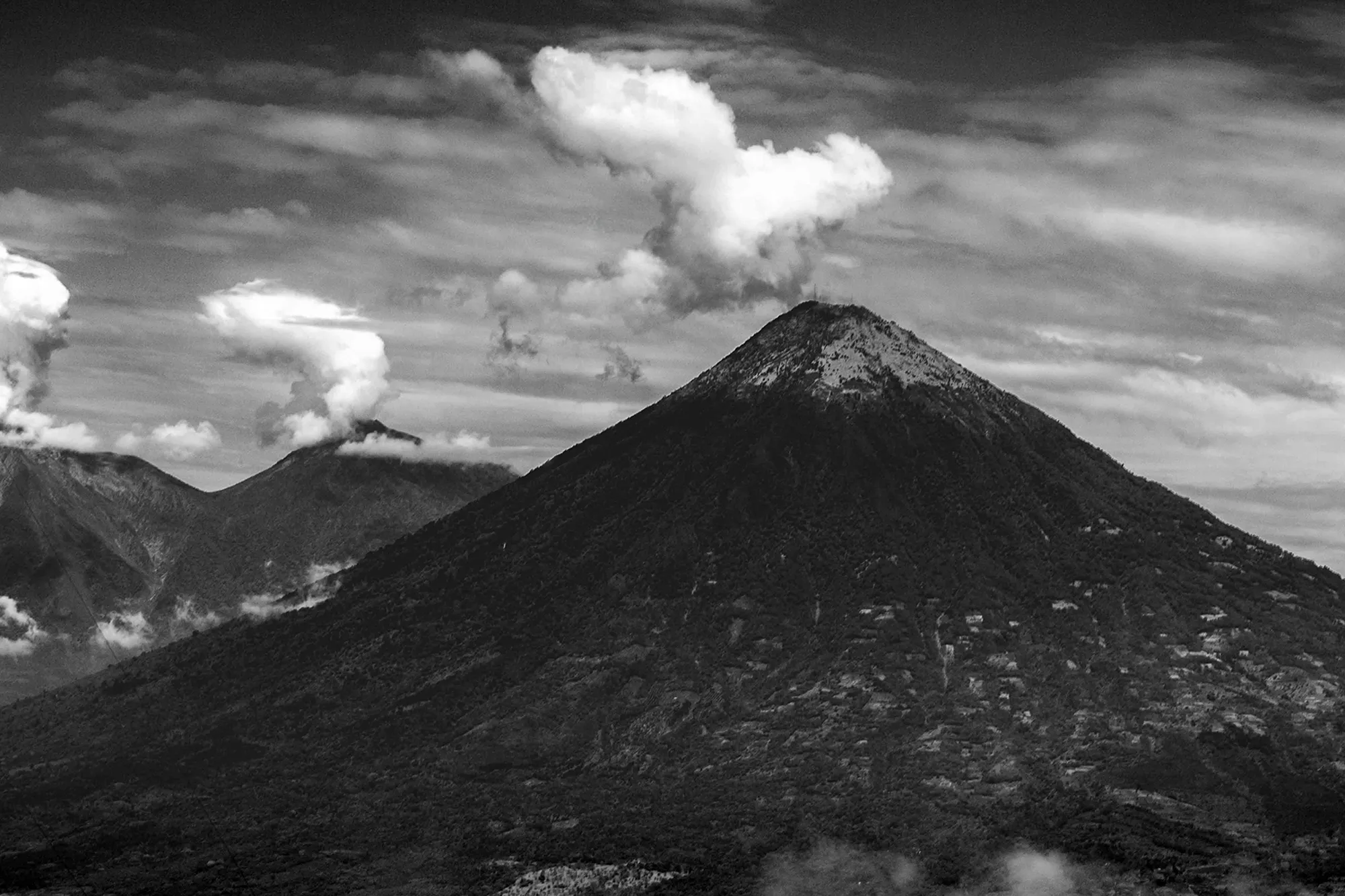 A steep conical volcano in black and white with a second, steaming cone rising behind it through the clouds